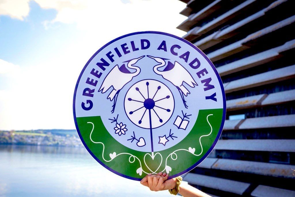 A school badge for Greenfield Academy being held up with a backdrop of the River Tay.