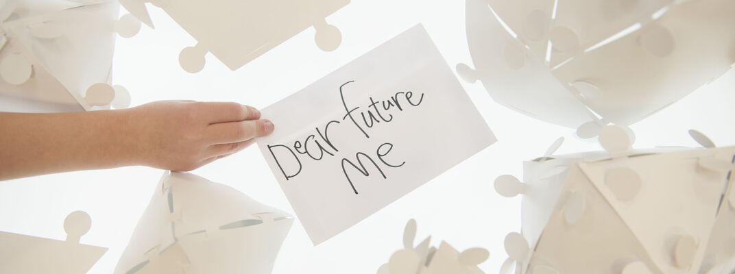 Someone holding a white piece of paper over a white light box with "Dear Future Me" on it scribbled in black pen.  The paper is surrounded by white geometric shapes made of paper.