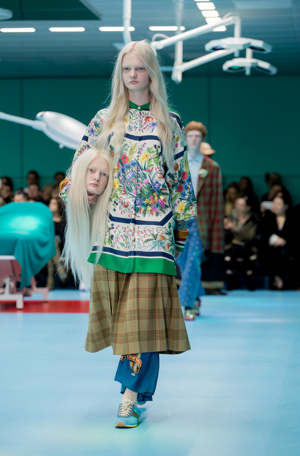 A model wearing colourful clothing walking down a runway holding a face head of themself.