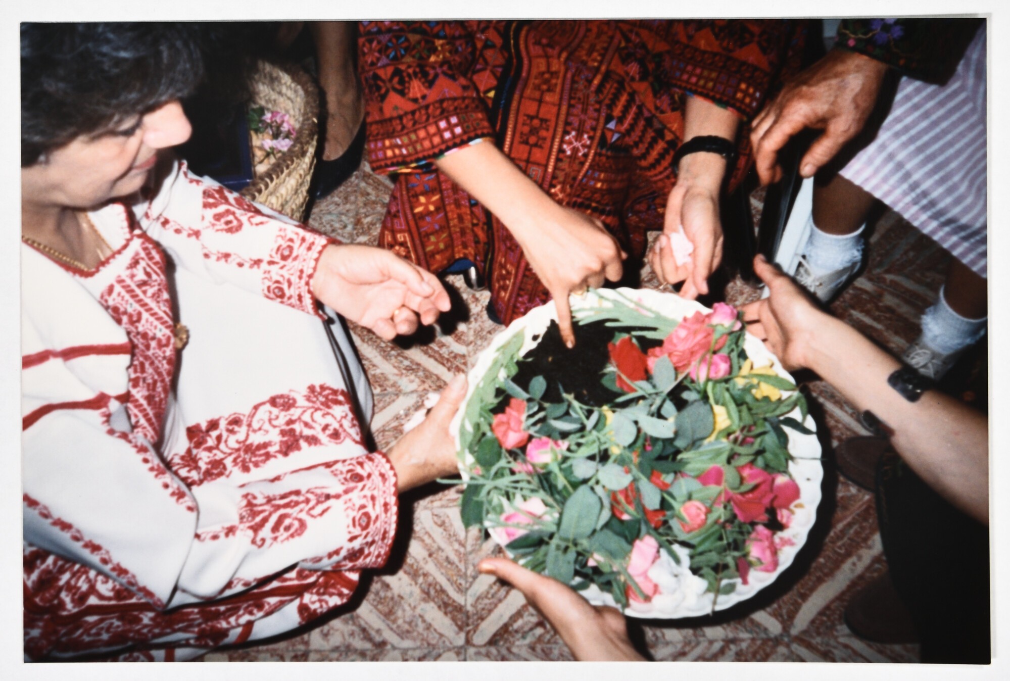 Women in traditional Palestinian dress gather around a bowl with soil and pink flowers.