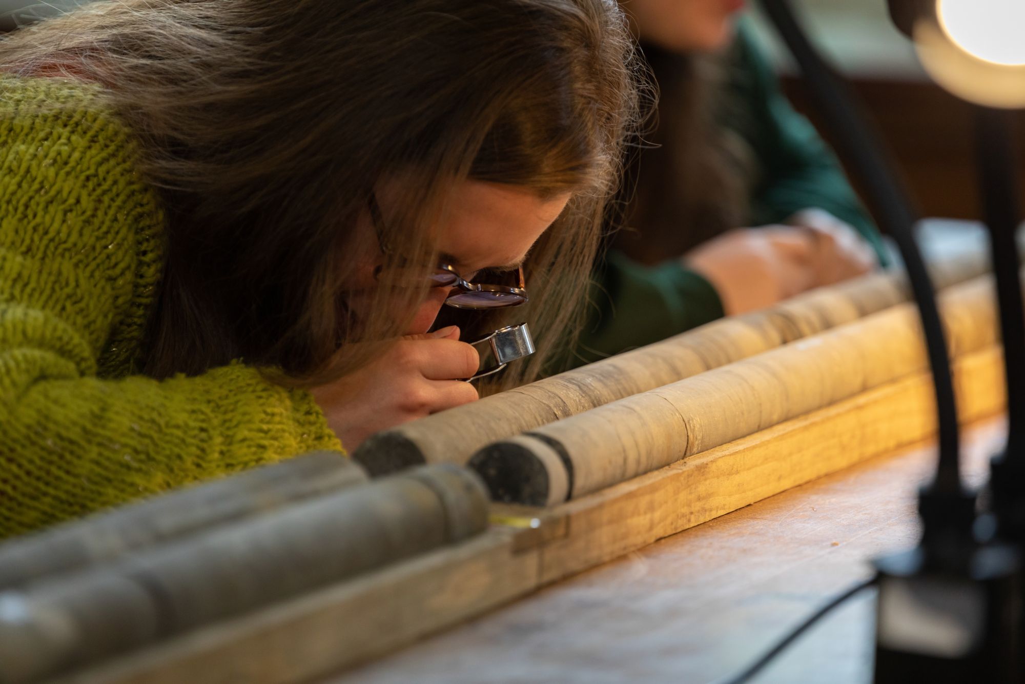 Person examining a wooden core sample with a magnifying glass on a table, under warm lighting.