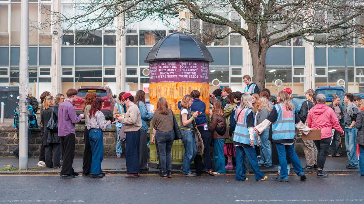 A group of people gathered around a large, umbrella-shaped art installation with text panels, on a city street with buildings in the background.
