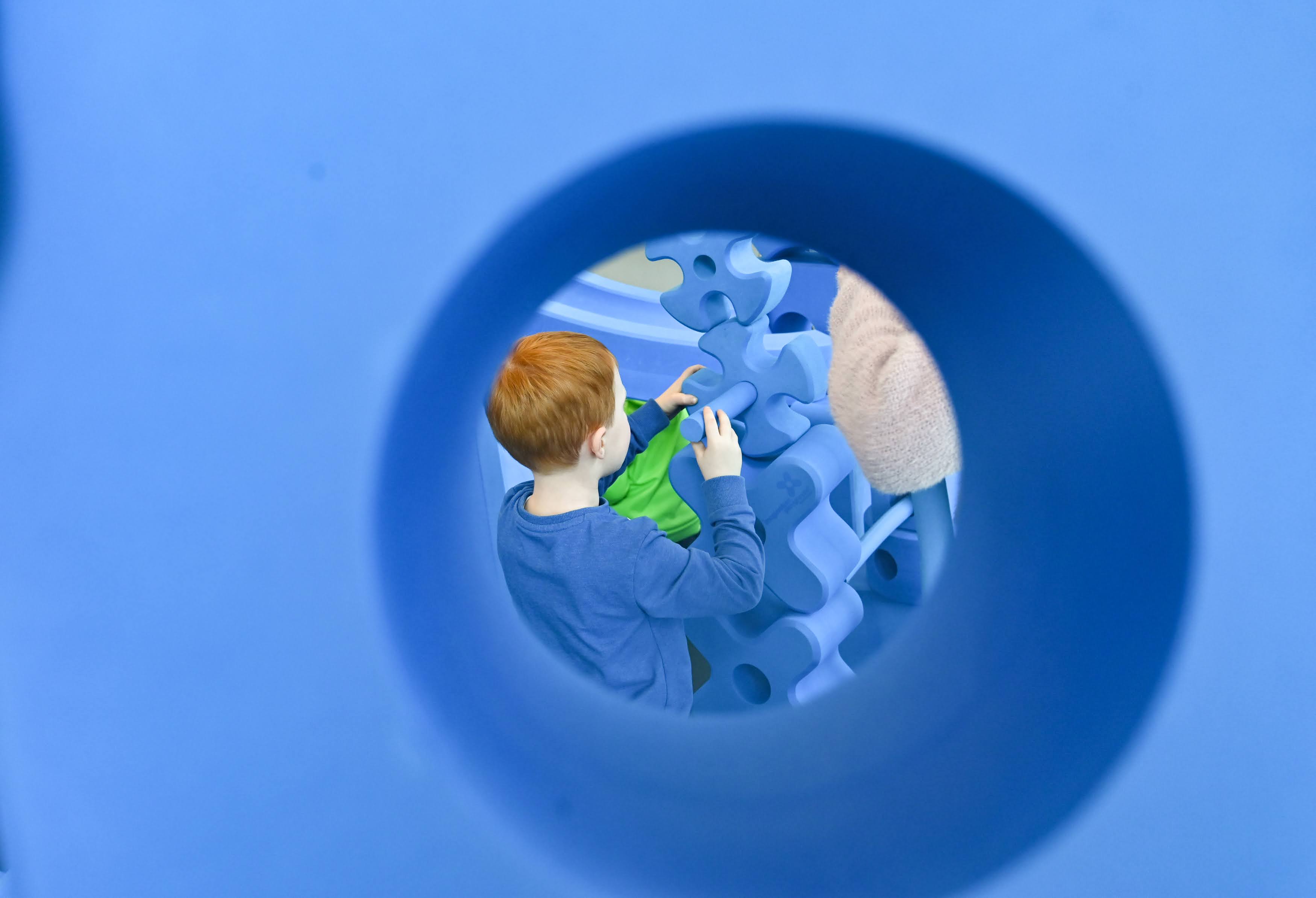 Child with red hair playing with large blue foam blocks, viewed through a circular opening.