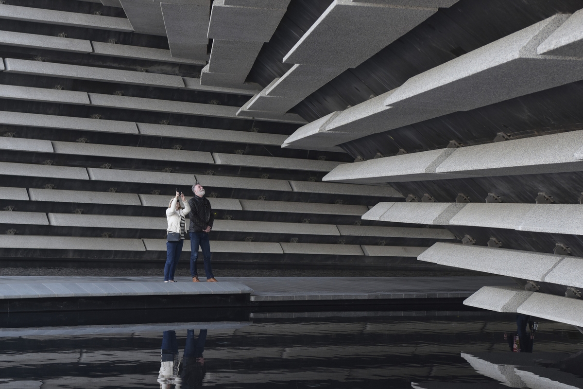 Two people stand beneath the V&A Dundee building with large, layered concrete slabs, next to a reflective water surface.