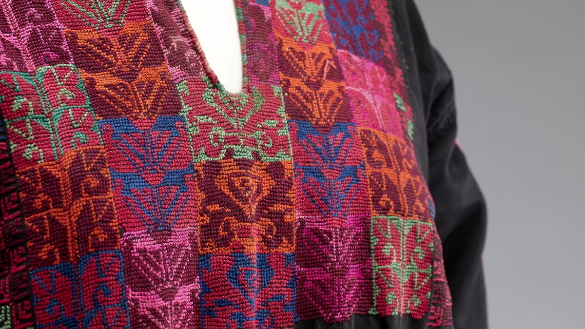 Close-up of a tatreez dress with colourful, intricately embroidered textile with geometric patterns in red, pink, green, and blue on a black fabric background.