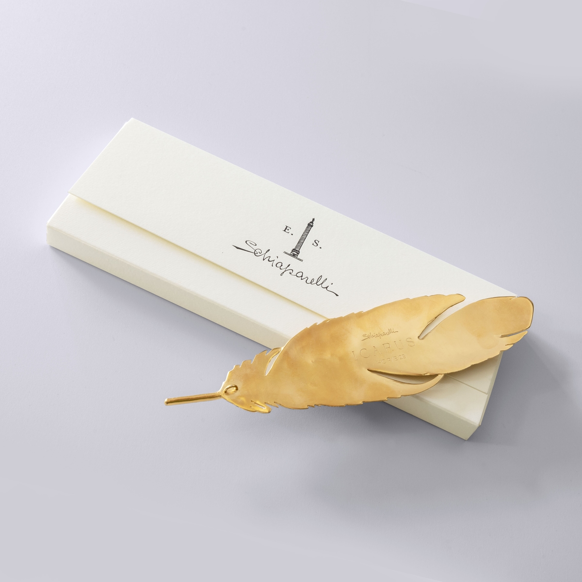 A gold feather is laid on top of a box which has the text 'Schiaparelli'