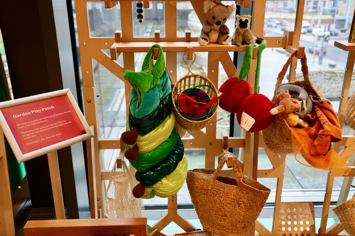 Colourful garden-themed play items hanging on a wooden display, including plush caterpillar and apples, near a window with an outdoor view.