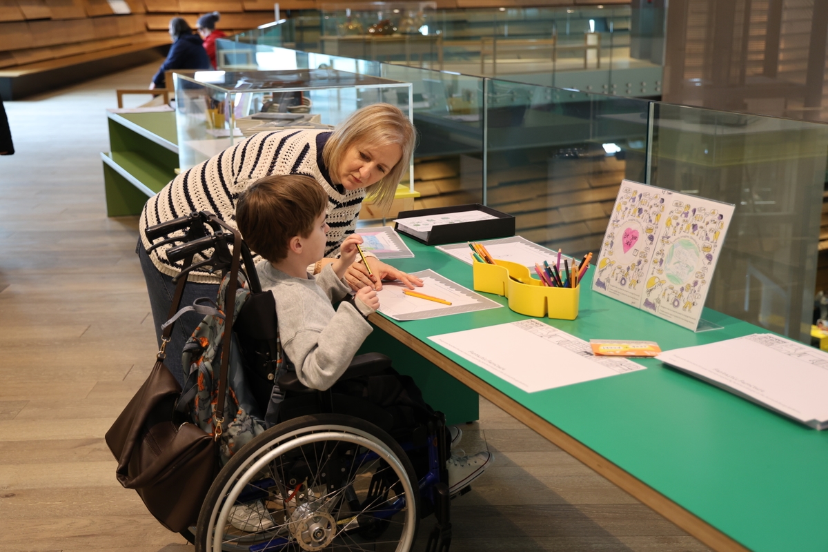 A person leaning over speaking to a child in a wheelchair as they draw on a green table.