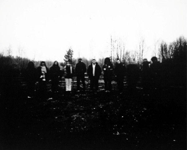 A group of people stand closely together in an open area with leafless trees, under a bright, overcast sky. The image is blurry and monochromatic.