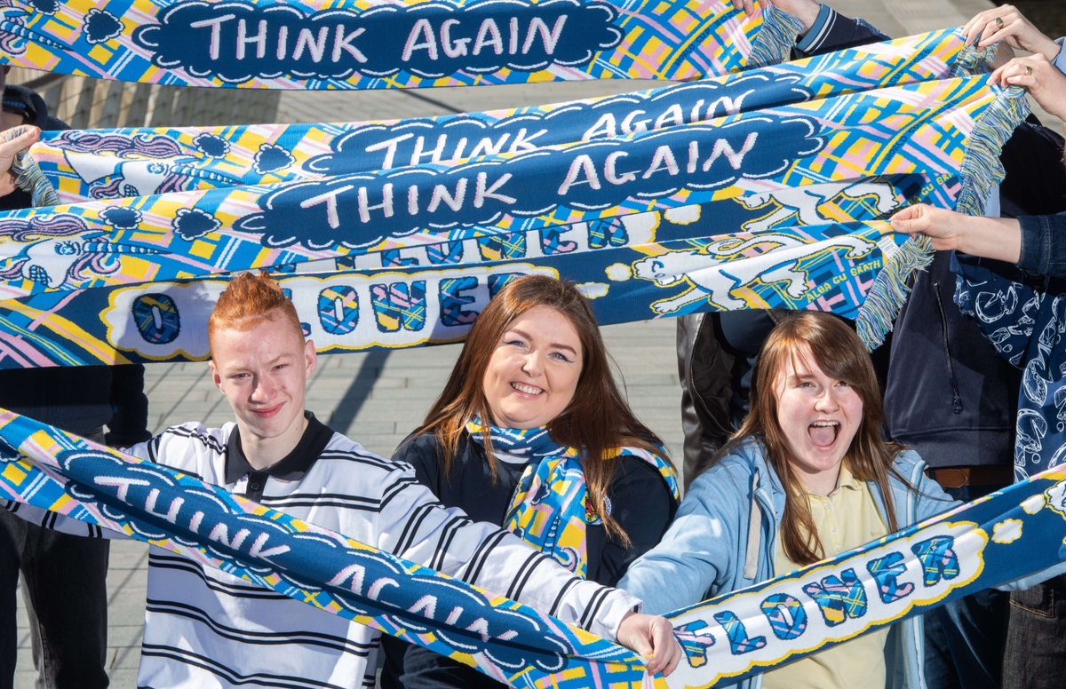 Three people hold colourful scarves that read "THINK AGAIN" and "FLOWER," smiling and cheering outdoors.
