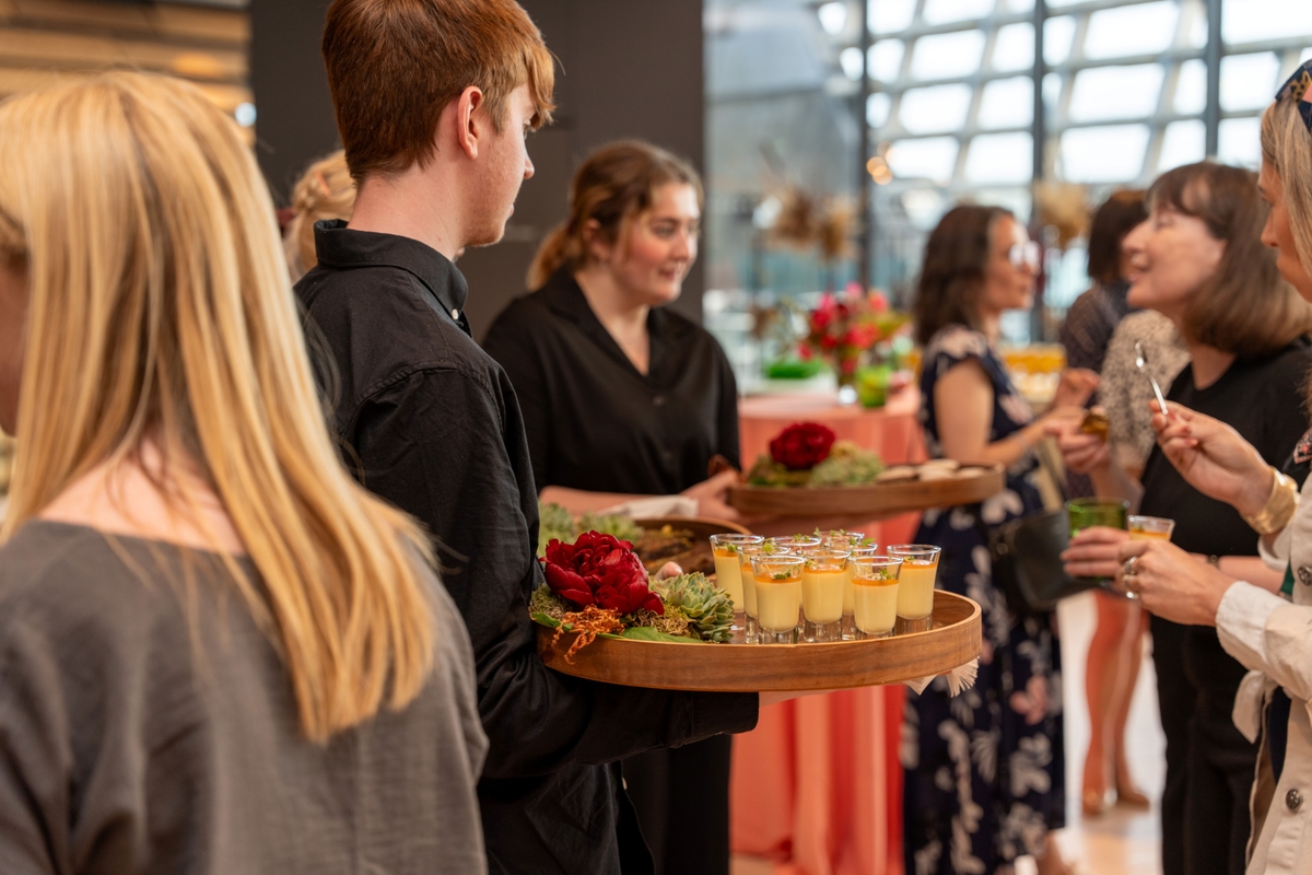 Servers in black attire offer trays of desserts at a lively event, with guests mingling and holding drinks in a modern, bright venue.