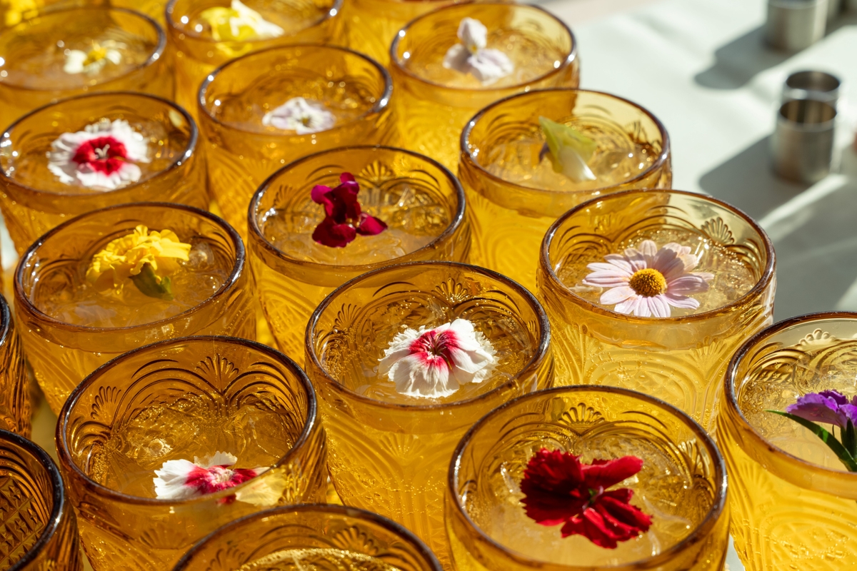 Rows of amber-coloured glasses filled with iced drinks, each topped with vibrant, decorative flowers, are arranged on a sunlit table.