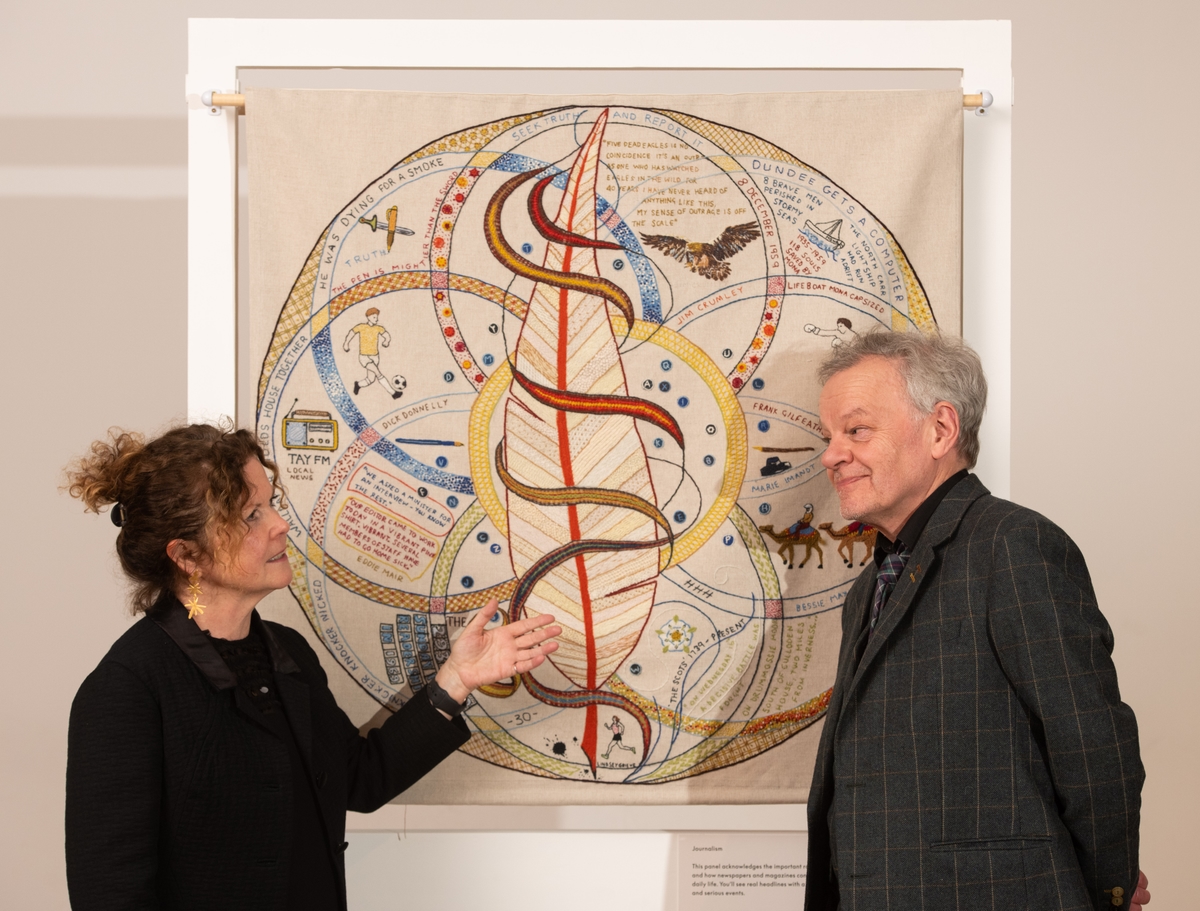 Two people standing beside a woven tapestry talking.