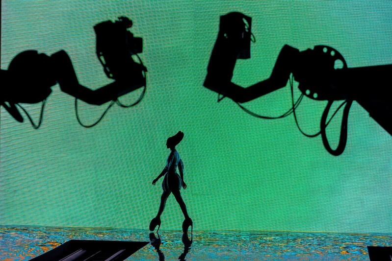 Silhouetted performer on stage with robotic arms overhead, set against a vibrant green background.