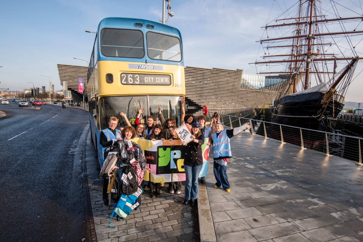 Group of people holding flags and banners in front of a vintage double-decker bus labled "263 City Centre," with a ship and V&A nearby.