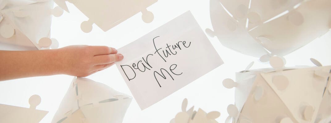 Someone holding a white piece of paper over a white light box with "Dear Future Me" on it scribbled in black pen.  The paper is surrounded by white geometric shapes made of paper.