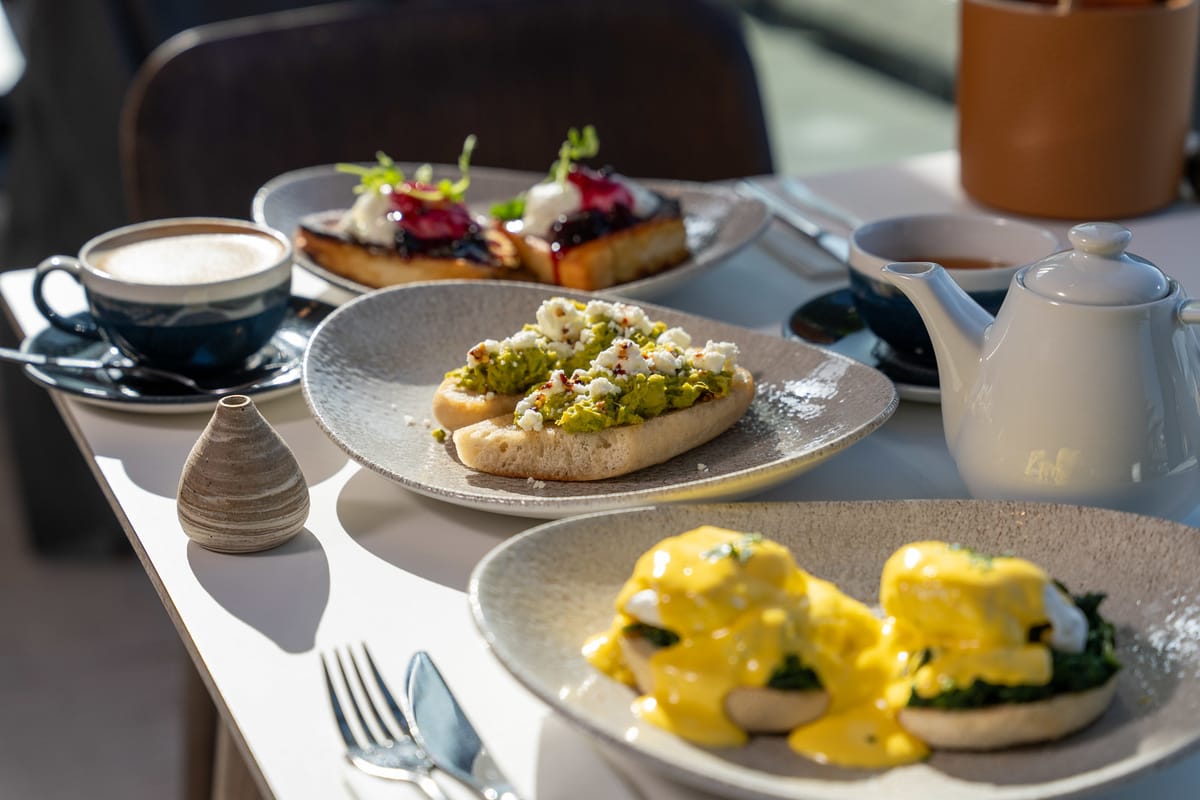 Breakfast spread with avocado toast, eggs benedict, French toast, cappuccino in a blue cup, and a white teapot on a sunlit table.