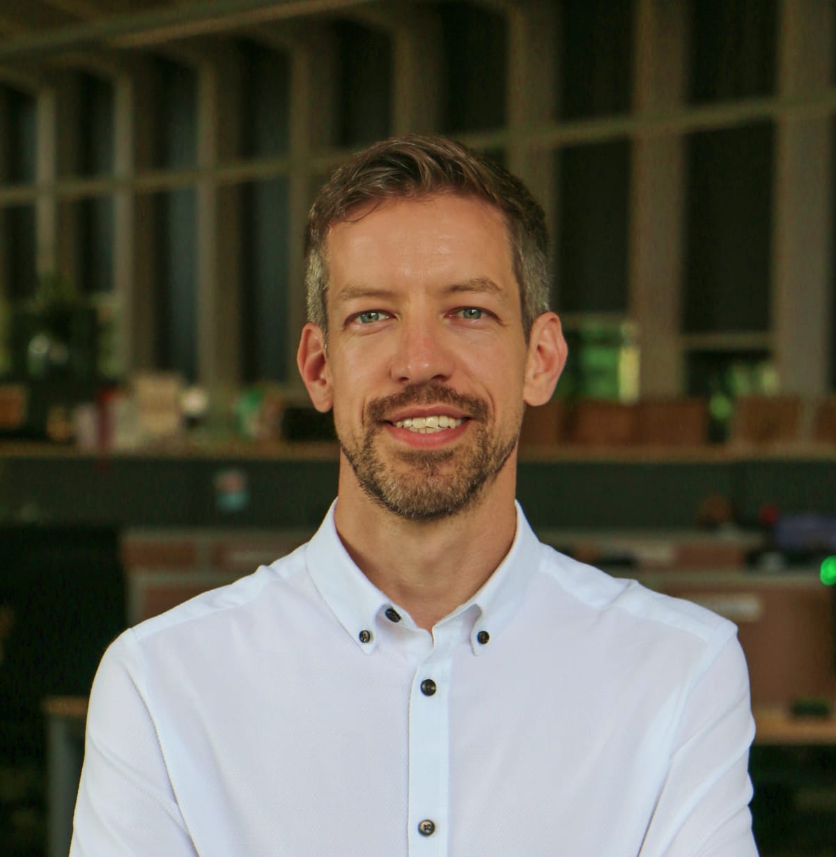 A man with short hair and a beard smiles, wearing a white shirt with black buttons, standing indoors with a blurred office background.