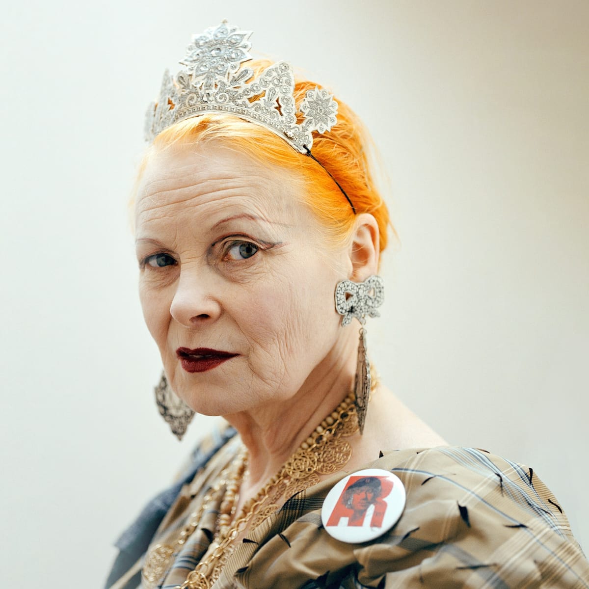 A woman with bright orange hair, wearing a detailed silver tiara, ornate earrings, and a patterned outfit with a button, gazes intently.
