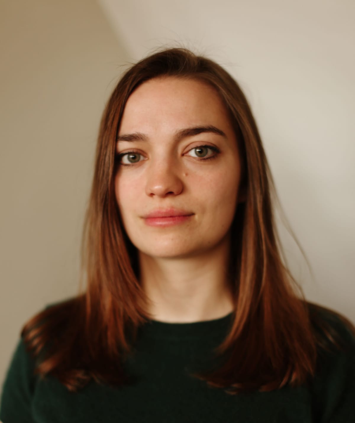 A person with long brown hair, wearing a dark green sweater, stands against a neutral background, looking directly at the camera.