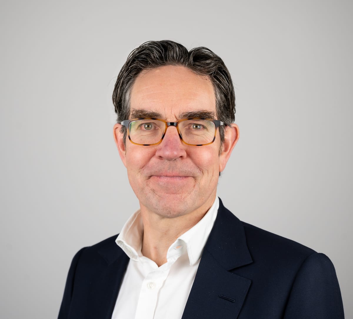 A man with glasses, wearing a white shirt and dark blazer, smiles slightly against a plain light background.