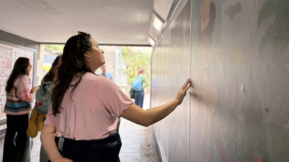 A woman in a pink shirt touches a mural on a tunnel wall, while others in the background engage in conversation.