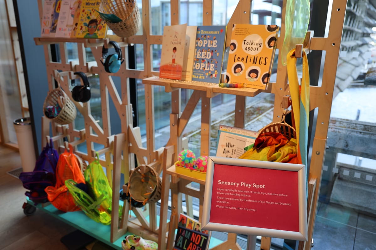 Sensory play area with colorful toys, books on empathy and feelings. Sign invites interaction, emphasising design and disability themes.