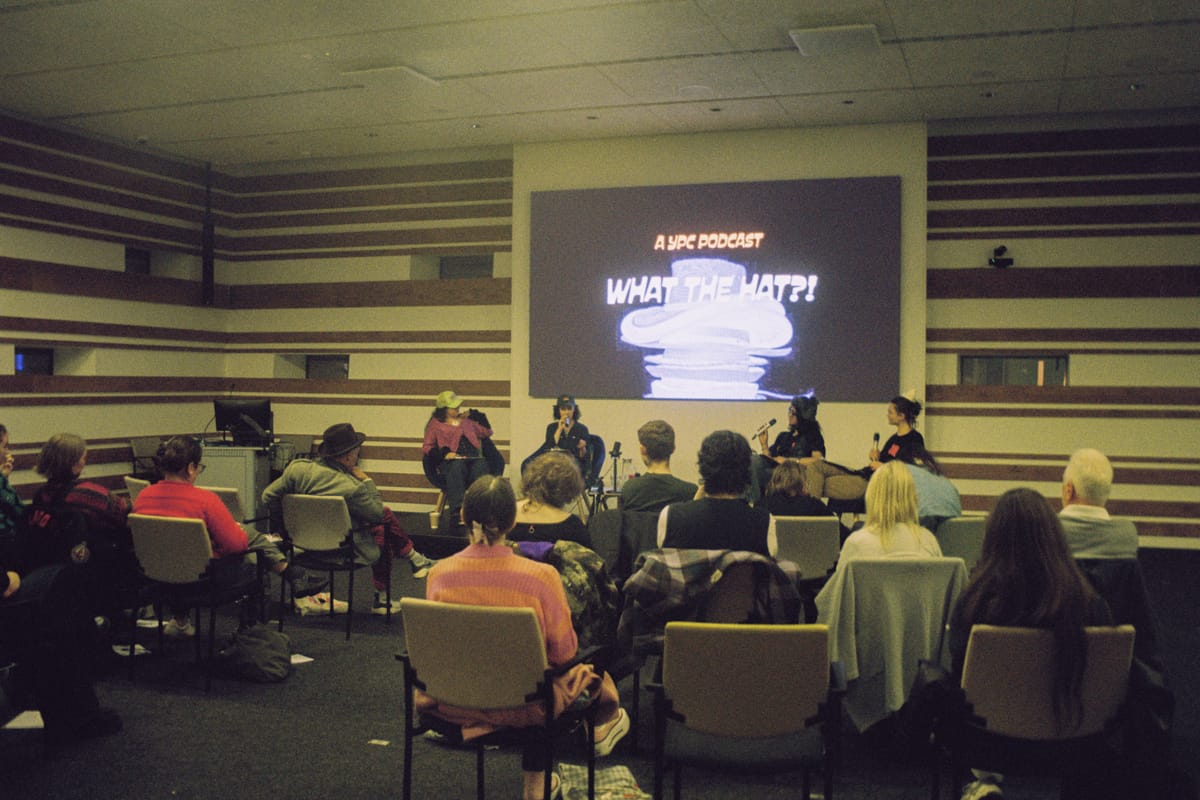 An auditorium filled with people seated looking towards a stage with four people seated. On the screen behind them reads "A YPC Podcast, What the Hat?!"