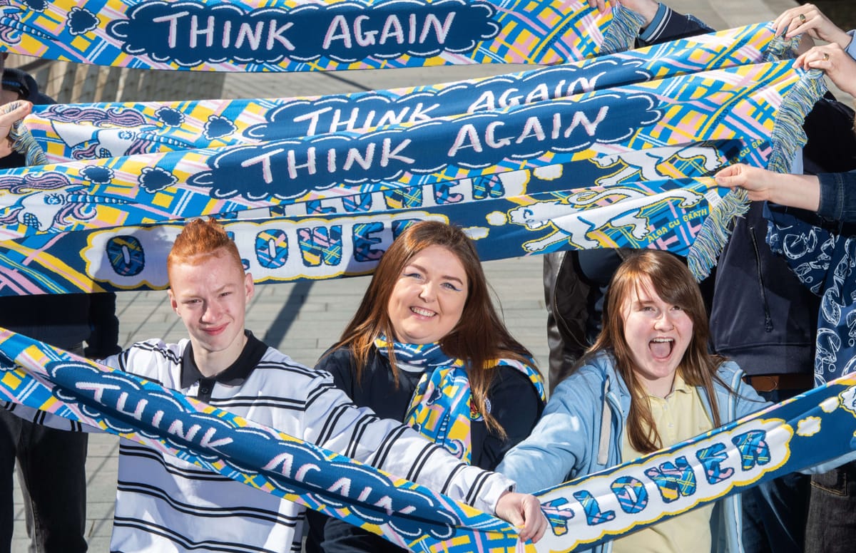 Three people hold colourful scarves that read "THINK AGAIN" and "FLOWER," smiling and cheering outdoors.