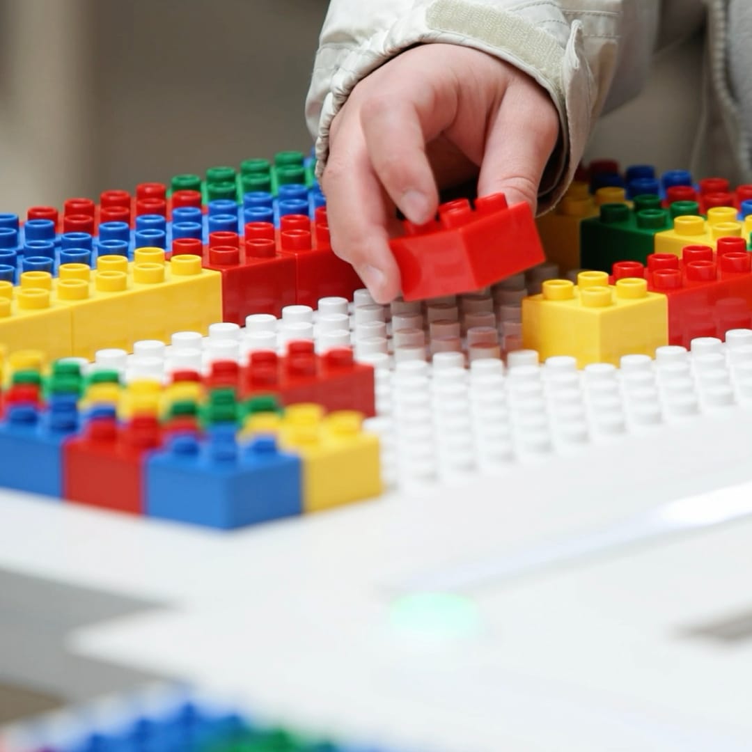 A hand building with colourful interlocking bricks on a white baseplate, featuring red, yellow, green, and blue pieces.