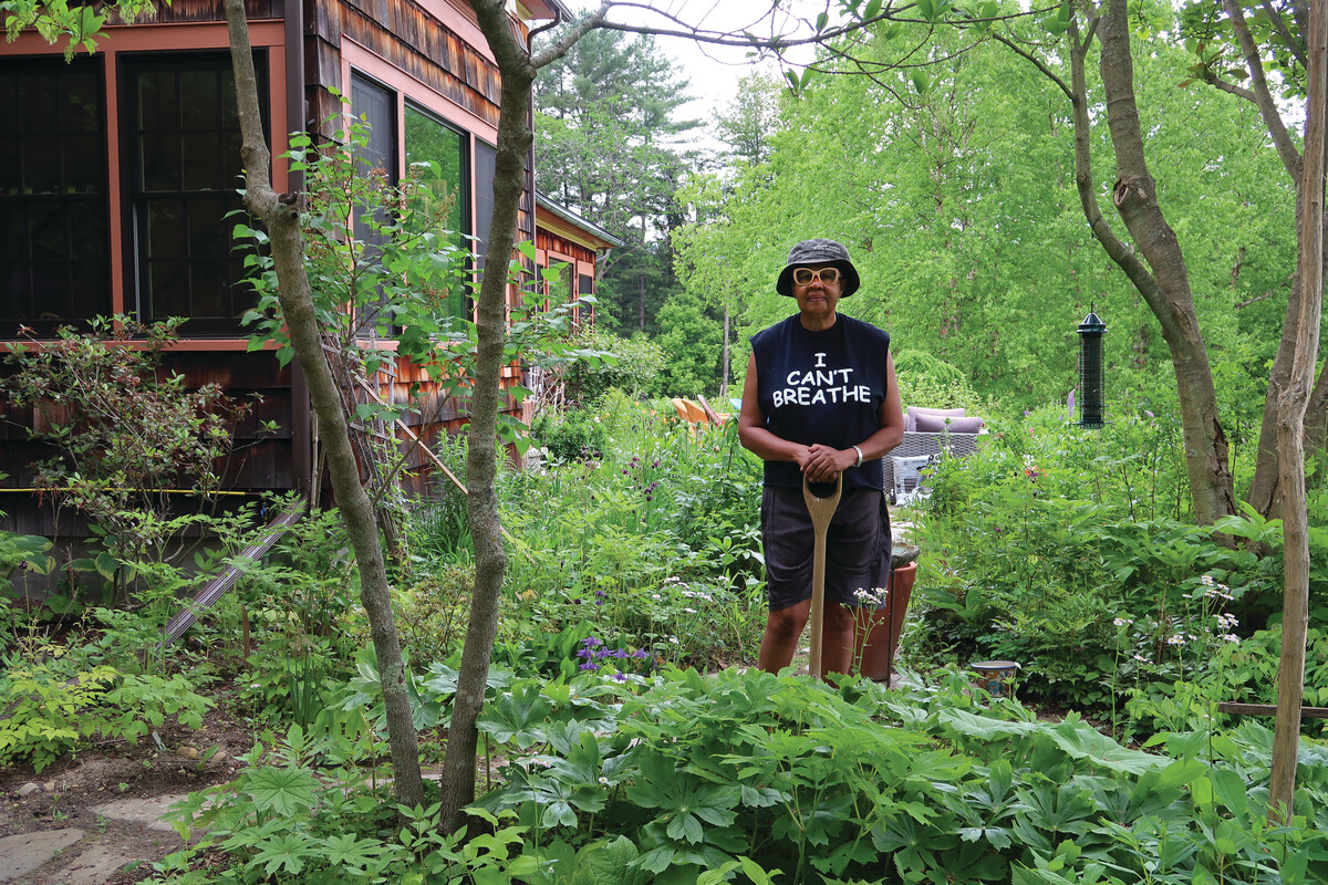 A person in a garden wearing a hat and a shirt with "I Can't Breathe" text, standing among lush greenery near a wooden house.