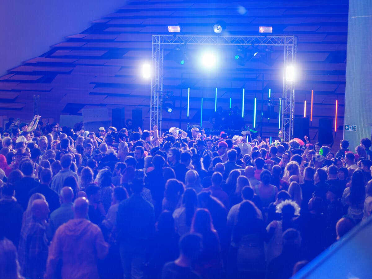 A large crowd gathers at an indoor music event with vibrant lighting and a DJ booth on stage, creating a lively and energetic atmosphere.