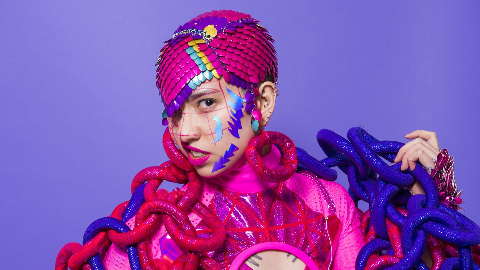 Person in vibrant pink and purple outfit with scale-patterned headpiece, colourful face paint, and large chain accessories on a purple background.