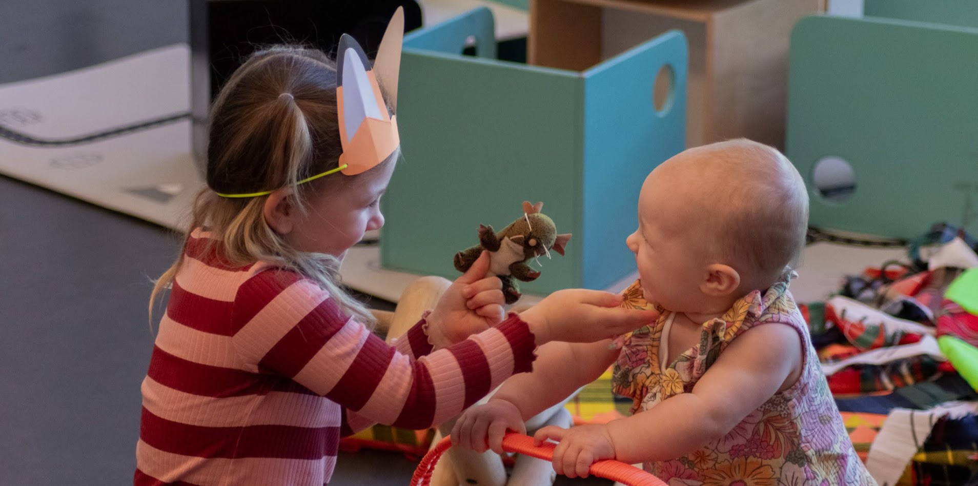 Two young children playing together. One child is wearing a striped top and is holding a toy out towards the baby sitting across from them