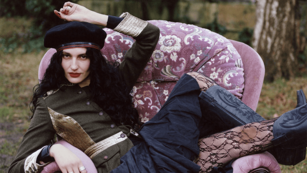 A woman with long curly hair reclines on a patterned armchair outdoors, wearing a military-style jacket, black lace leggings, and boots.
