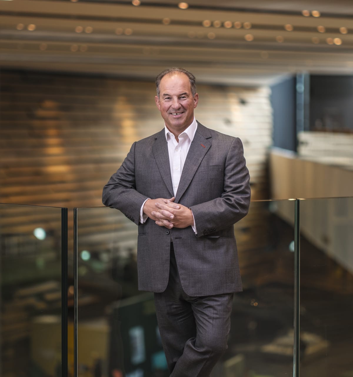 Man in a gray suit stands confidently by a glass railing indoors, with a blurred modern architectural background.