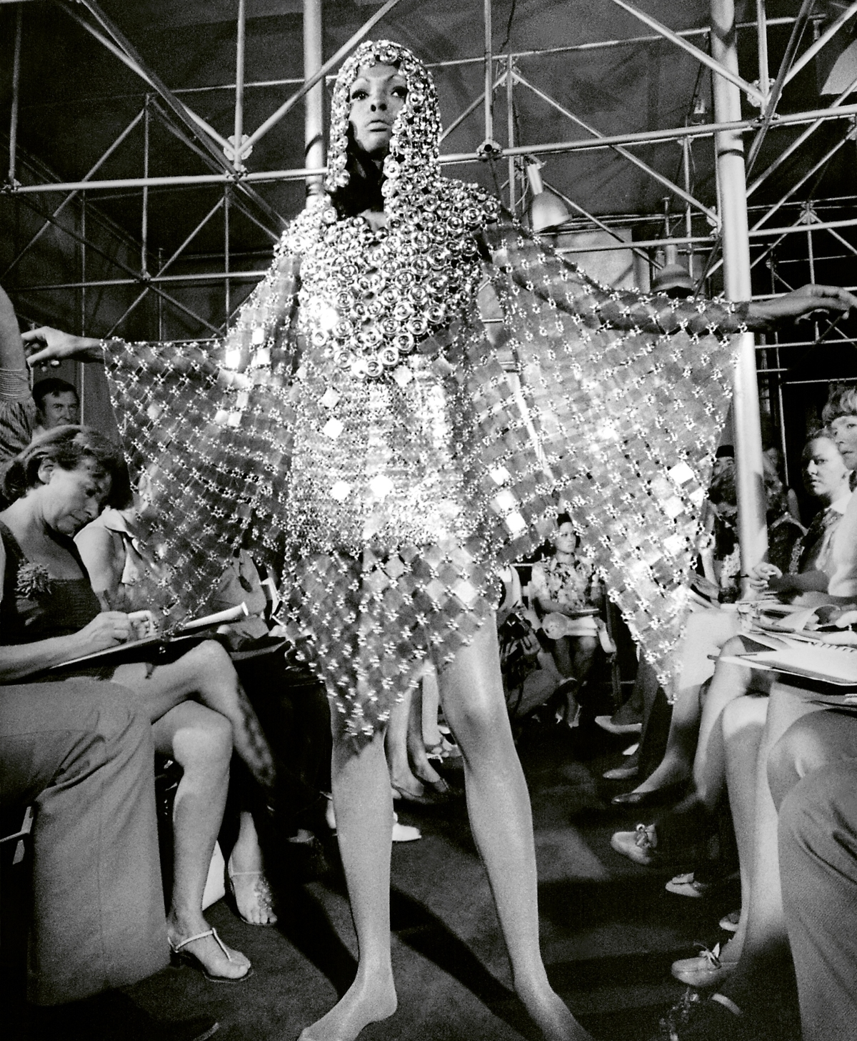 A black and white image of a model wearing a metal dress, people are seated down the runway.