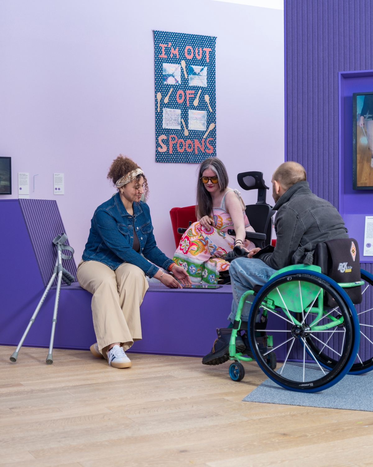 A group of people interacting in a colourful room with purple accents. One person is seated in a wheelchair with green wheels, and another is looking at a tablet. A tapestry on the wall behind them reads, "I'M OUT OF SPOONS".