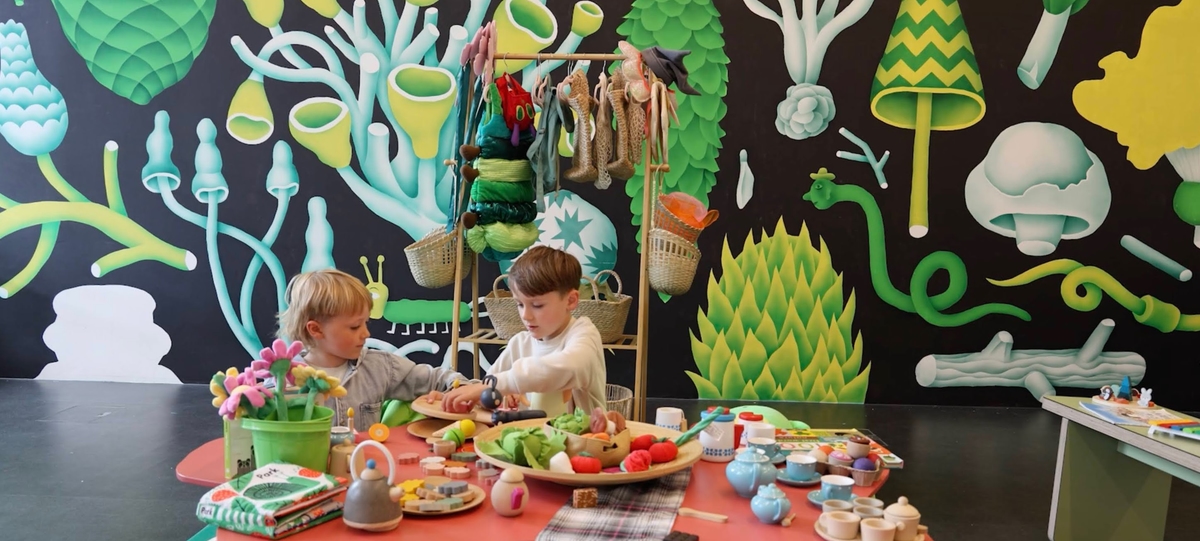 Two children sit at a decorated table with toy food, and tea sets, in front of a colourful mural featuring plants