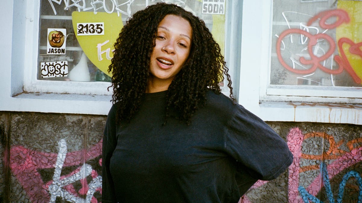 A woman with curly hair and a nose ring stands in front of a graffiti-covered wall, smiling with a relaxed expression.