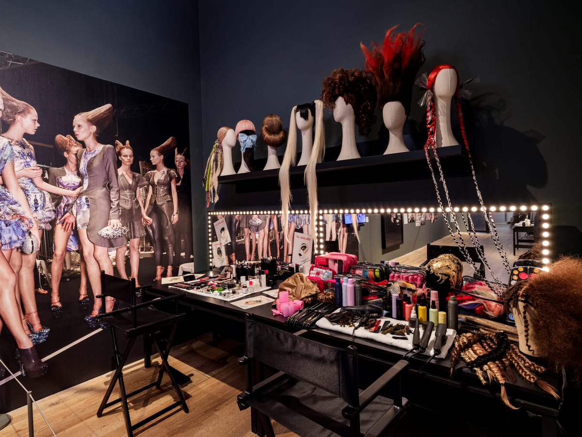 A makeup station with wigs on mannequins, beauty products, and hair tools, surrounded by a wall poster of models with elaborate hairstyles.