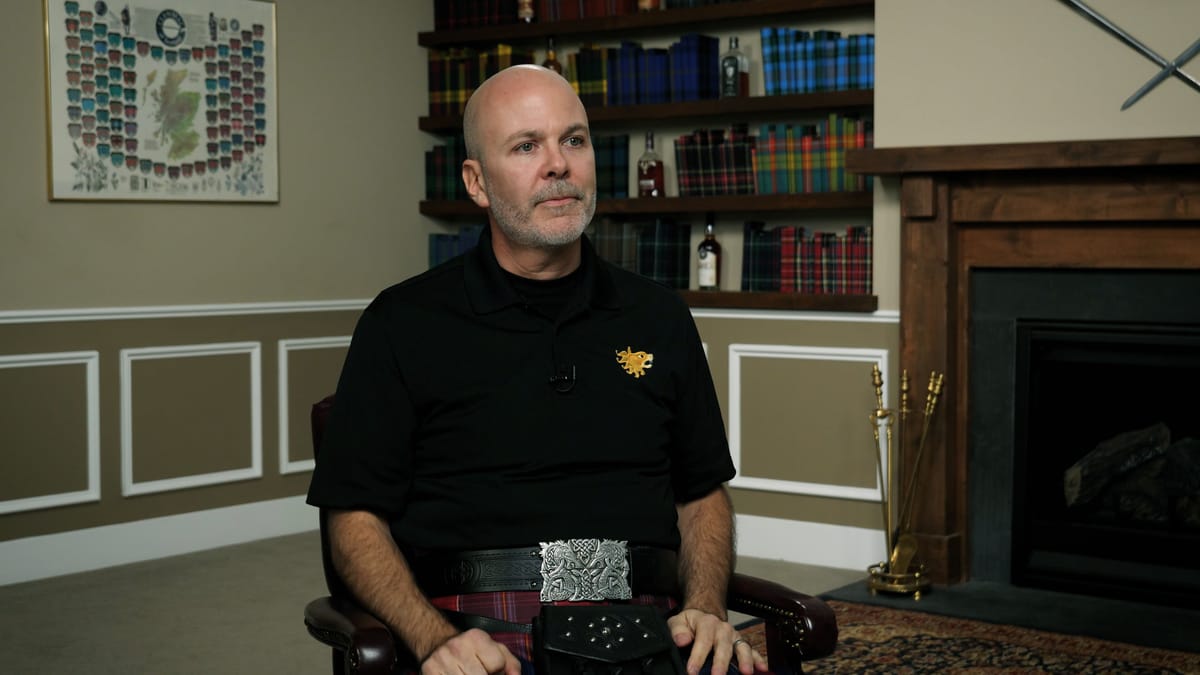 A man sitting in front of a fireplace wearing a black jumper and kilt.