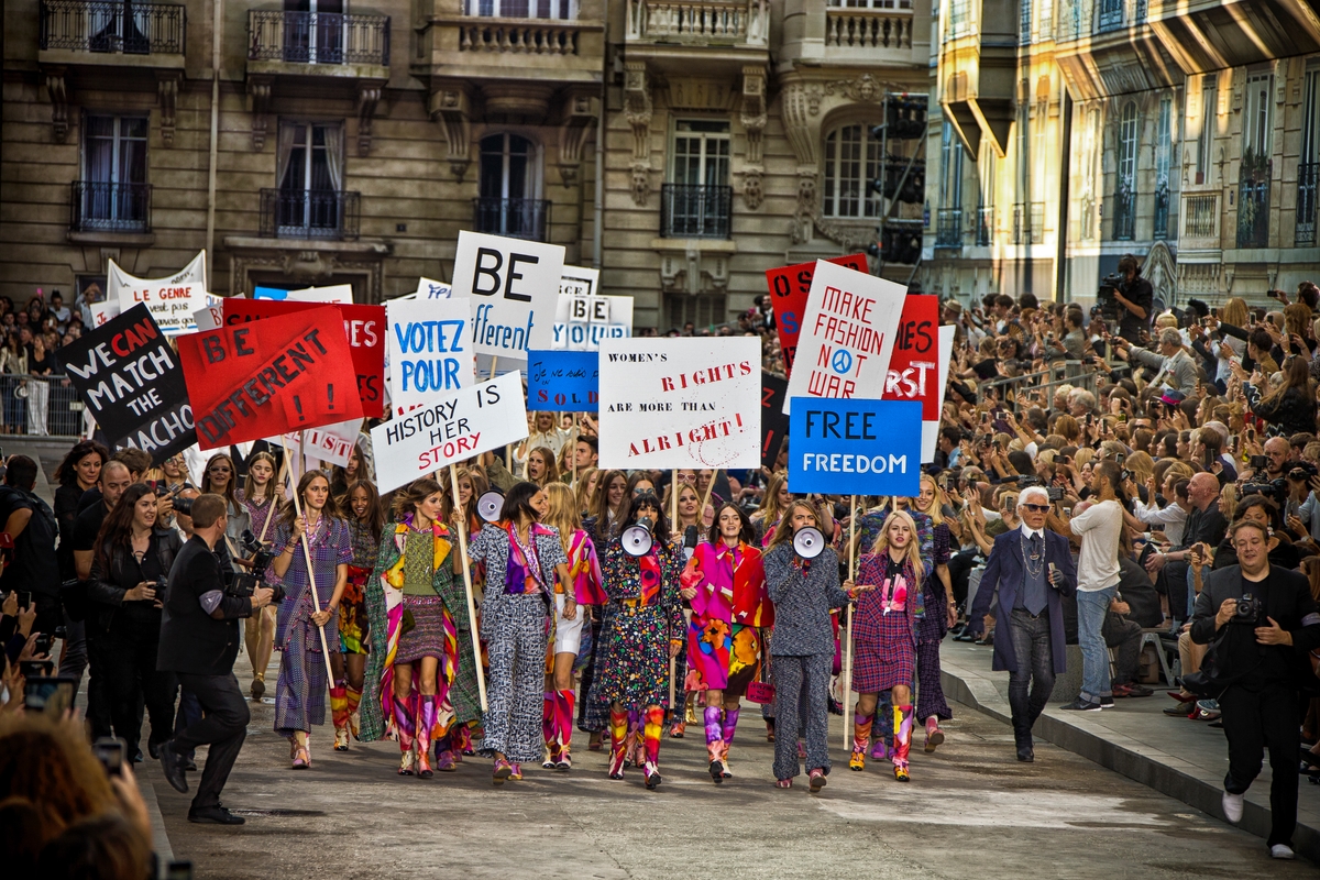 A group of models on stage which is fashioned as a street, they're holding signs and protesting.