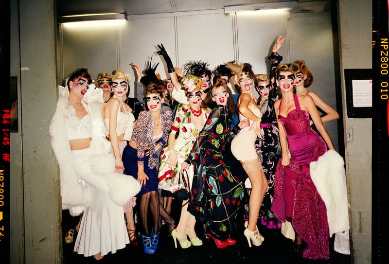 A group of fashion models in eclectic, colourful costumes and exaggerated makeup pose energetically in a hallway, celebrating in a playful atmosphere.